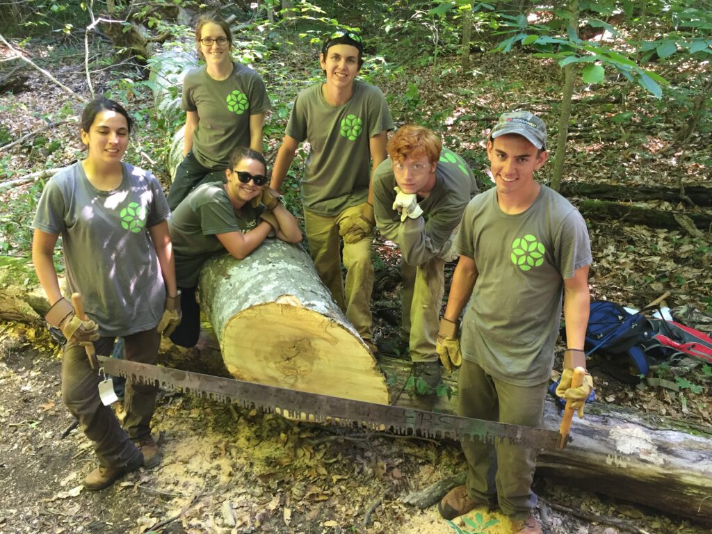 A handful of EcoCorps workers stand with a large felled tree and the tree saw used in trail clearing.