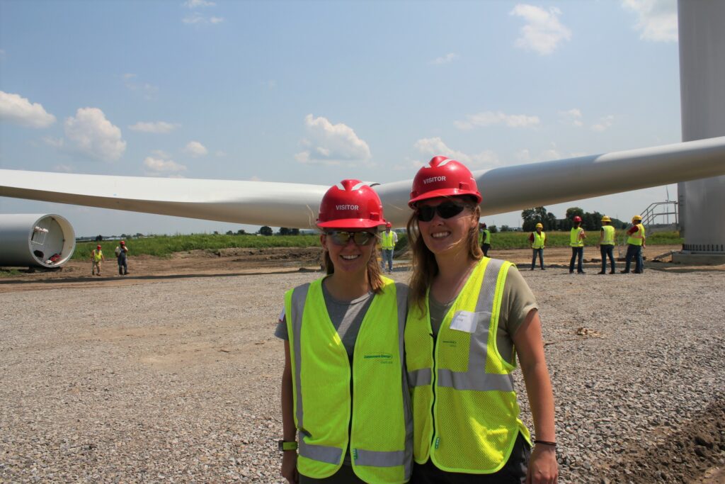 Two women stand in front of pre-assembled wind turbine showing the large scale of the blade compared to people.