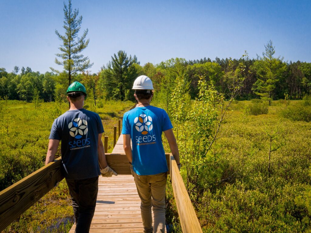 Two people in hard hats and SEEDS shirts are shown from behind while constructing part of the North Country Trail.