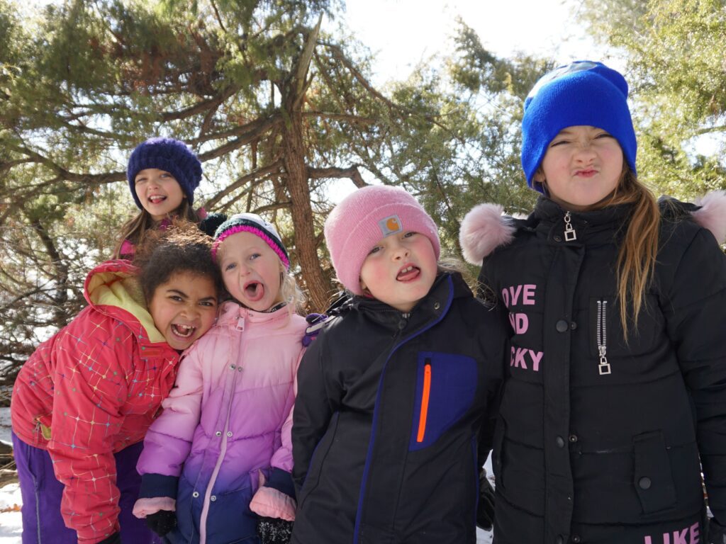 A group of students in winter coats and hats smiles and poses together in a forest.