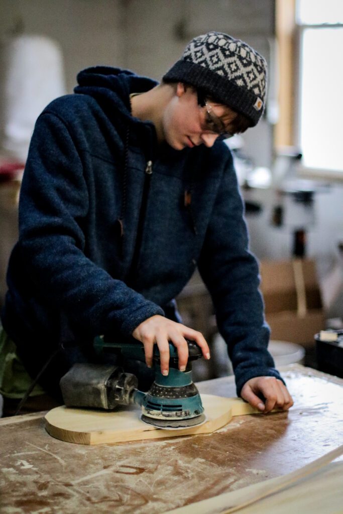 A carpentry student in a hoodie and a beanie uses a hand-sander to sand a wood project in a workshop.