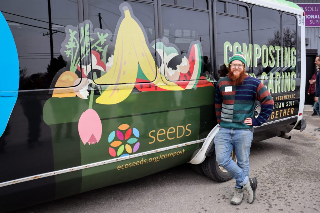 A city bus with a cartoonish compost bin featuring SEEDS logo is overflowing with giant compostable materials incl. banana peel, apple cores, carrot stems, flowers and fruit rinds